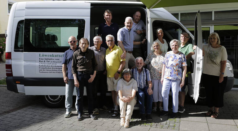 Eine Gruppe von Menschen steht und sitzt lächelnd vor einem weißen Minibus mit offener Tür.