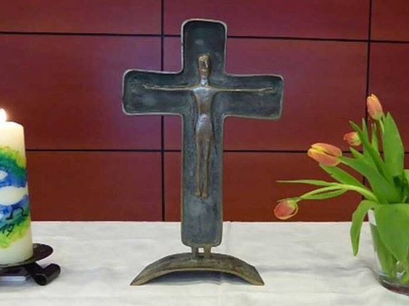 Auf einem Tisch stehen eine Kerze mit buntem Muster, ein metallisches Kreuz mit einer Figur darauf und eine Vase mit orangefarbenen Tulpen.