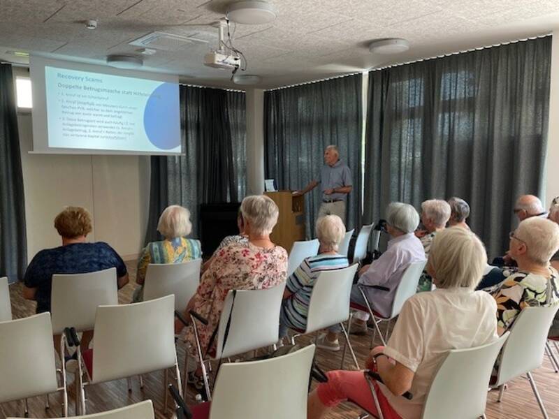Eine Gruppe älterer Menschen sitzt in einem Raum und hört einem Vortrag zu, der mit einer Präsentation auf einer Leinwand begleitet wird.