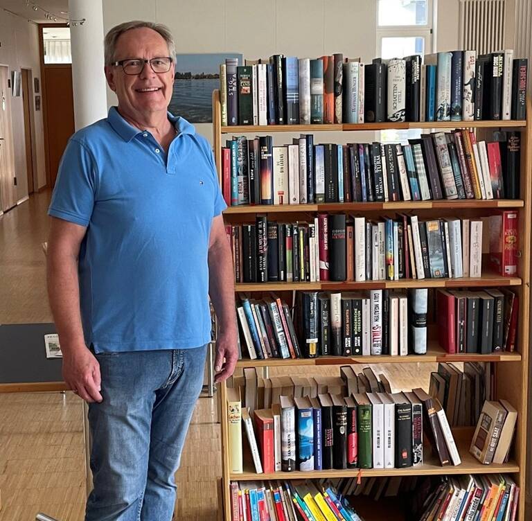 Ein Mann in einem blauen Polohemd steht lächelnd neben einem Bücherregal, das mit zahlreichen Büchern gefüllt ist.