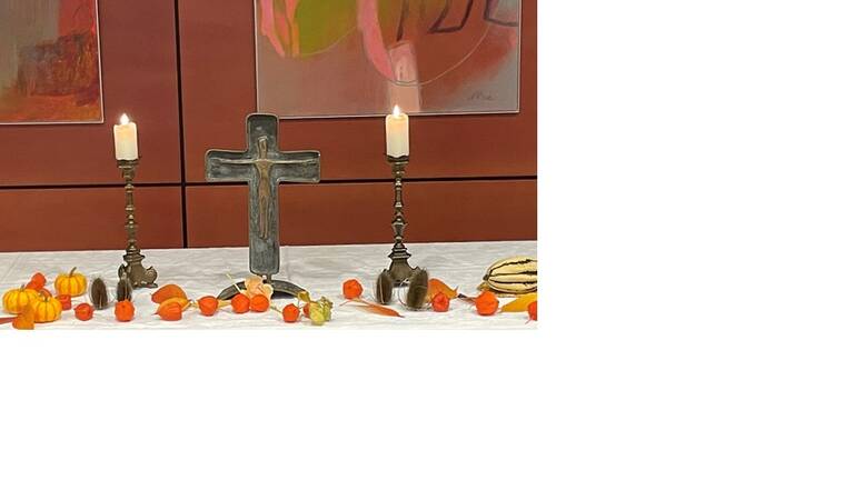 Auf einem Tisch stehen zwei Kerzenleuchter mit brennenden Kerzen, ein Metallkreuz in der Mitte und herbstliche Dekoration mit kleinen Kürbissen und Physalis.
